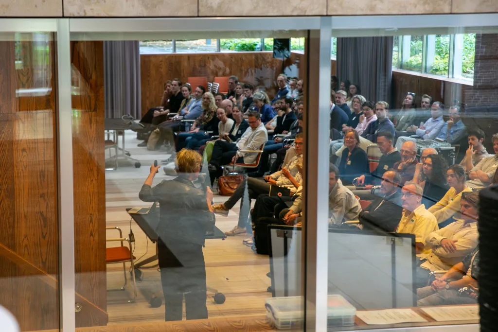 Seminarzaal van buiten gefotografeerd waar een publiek wordt toegesproken door een spreker