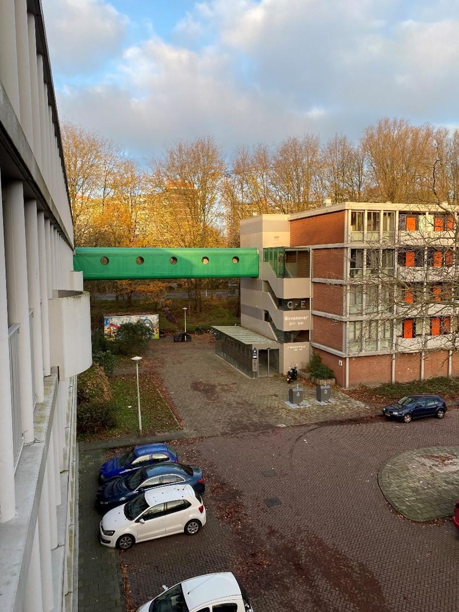 Een stedelijk landschap met twee flatgebouwen die een weg met parkeerplaats omringen met geparkeerde auto’s.